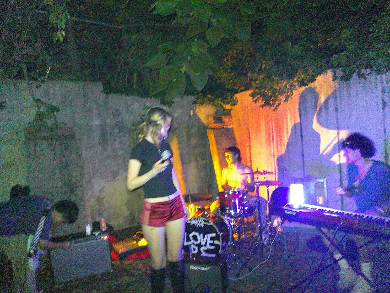 A campus house show with a singer, drummer, and keyboardist playing under colorful backyard lights
